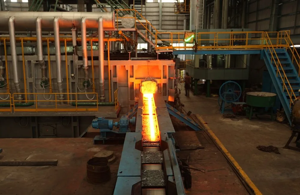A glowing hot steel billet leaving a reheat furnace onto a roller conveyor in a rolling mill